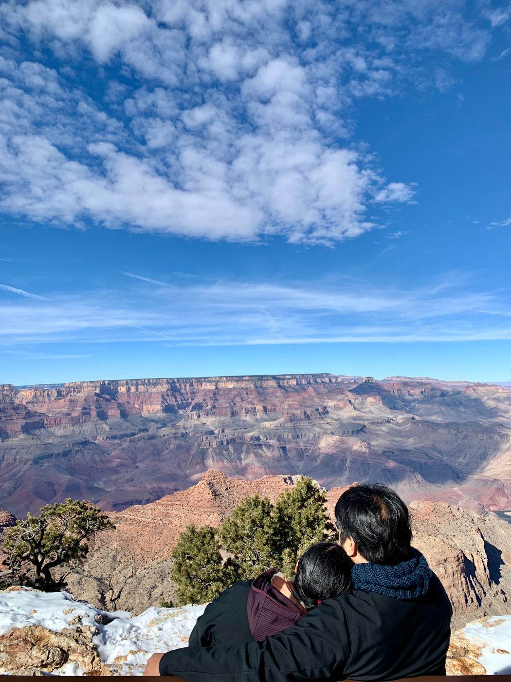 Two travellers wrapped up together on the rim of the Grand Canyon, looking out over the layered red cliffs under a wide blue sky